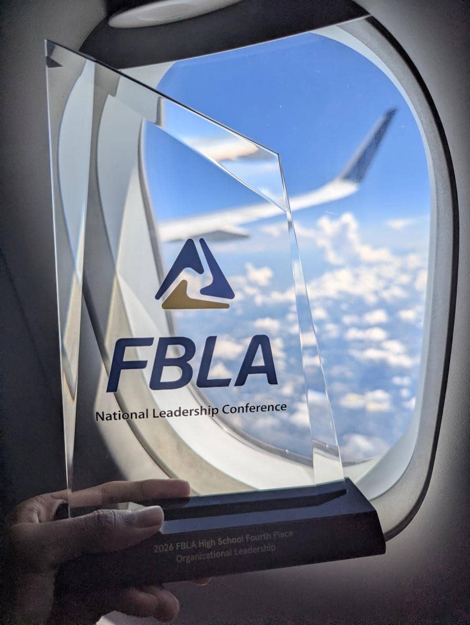 The FBLA glass award engraved 2026 FBLA High School Fourth Place Organizational Leadership, held against an aeroplane window above the clouds.