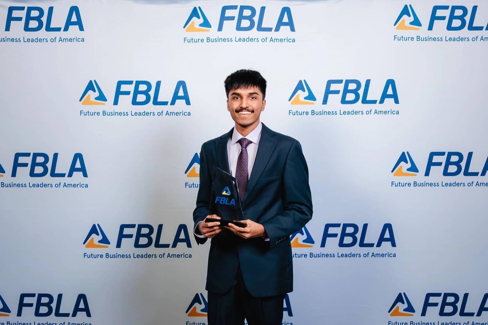 Sai Amartya in a suit holding the FBLA National Leadership Conference glass award in front of a step and repeat FBLA backdrop.